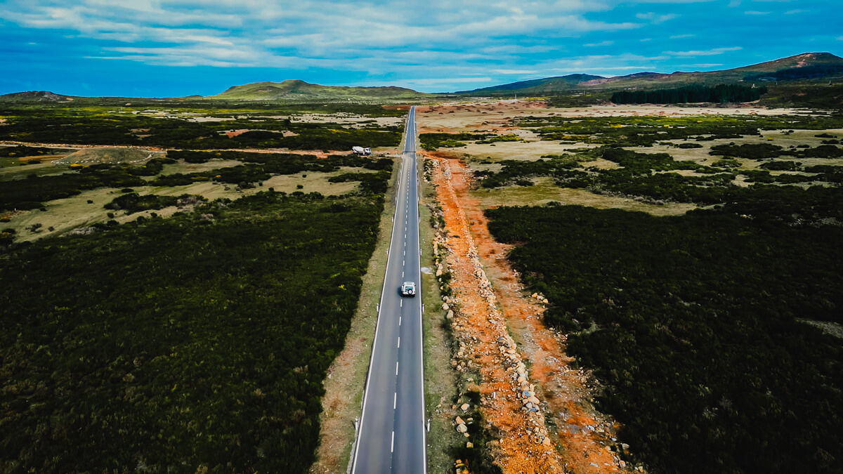 Drone shot of a jeep driving a straight road into the horizon with nature around 